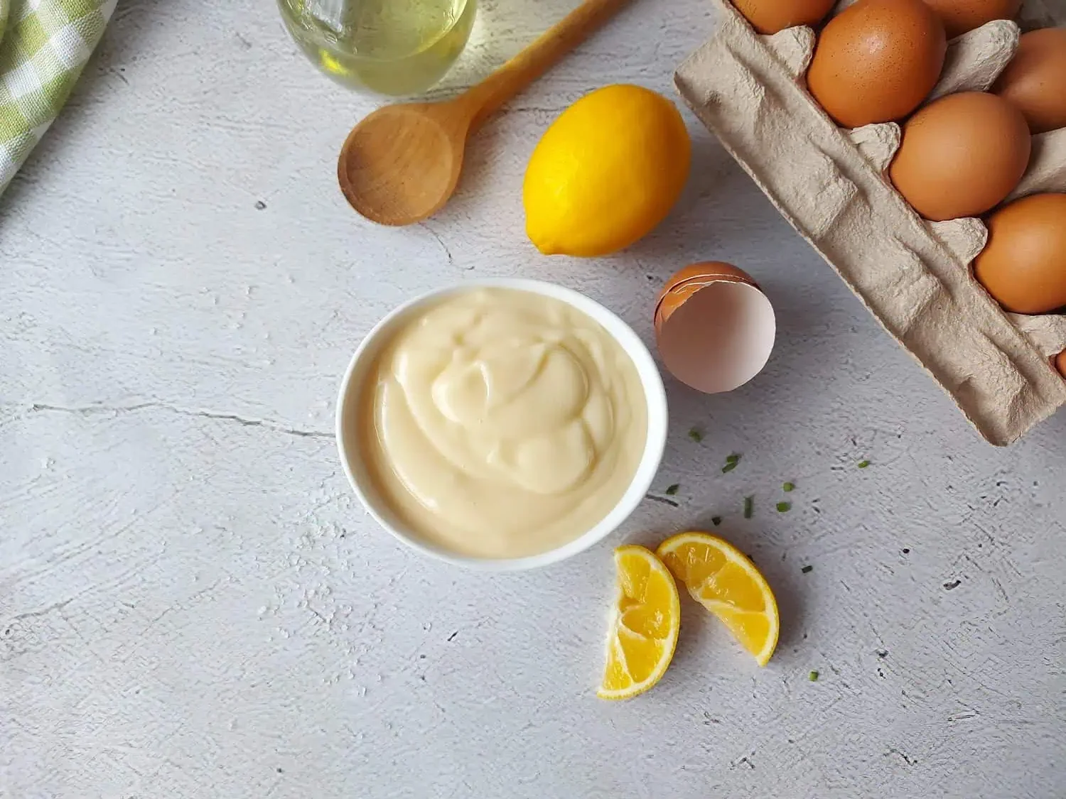 Mayonnaise sauce next to a lemon, egg shell, wooden spoon and oil