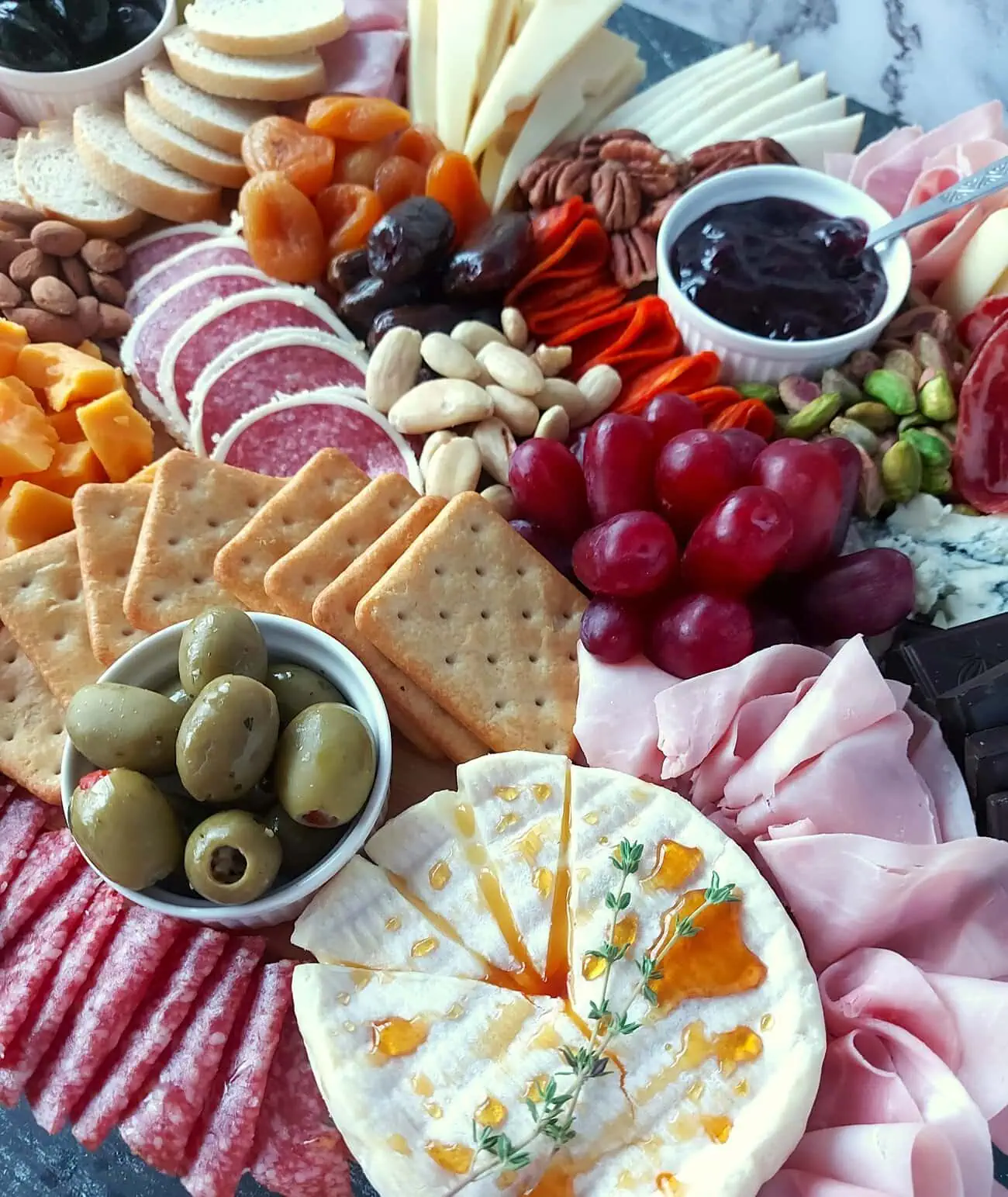 close-up of a brie cheese wheel drizzled with honey and a thyme sprig in the forefront, next to salami, ham, green olives, crackers, red grapes, pistachio, jam, and nuts
