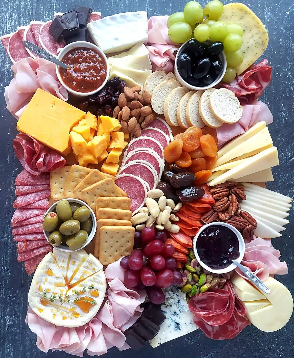 overtop image of a display of cheeses, meats, grapes, crackers, nuts, jams, fresh and dried fruit, olives and nuts