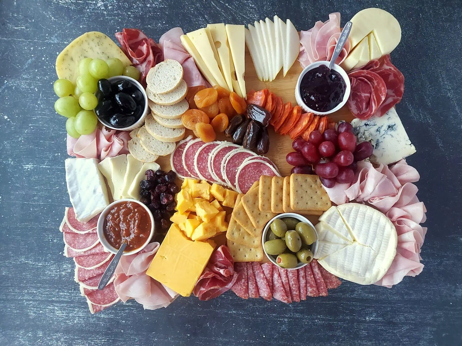wooden board with cheese, meats, crackers, sliced bread, grapes, jams, olives and dried fruits