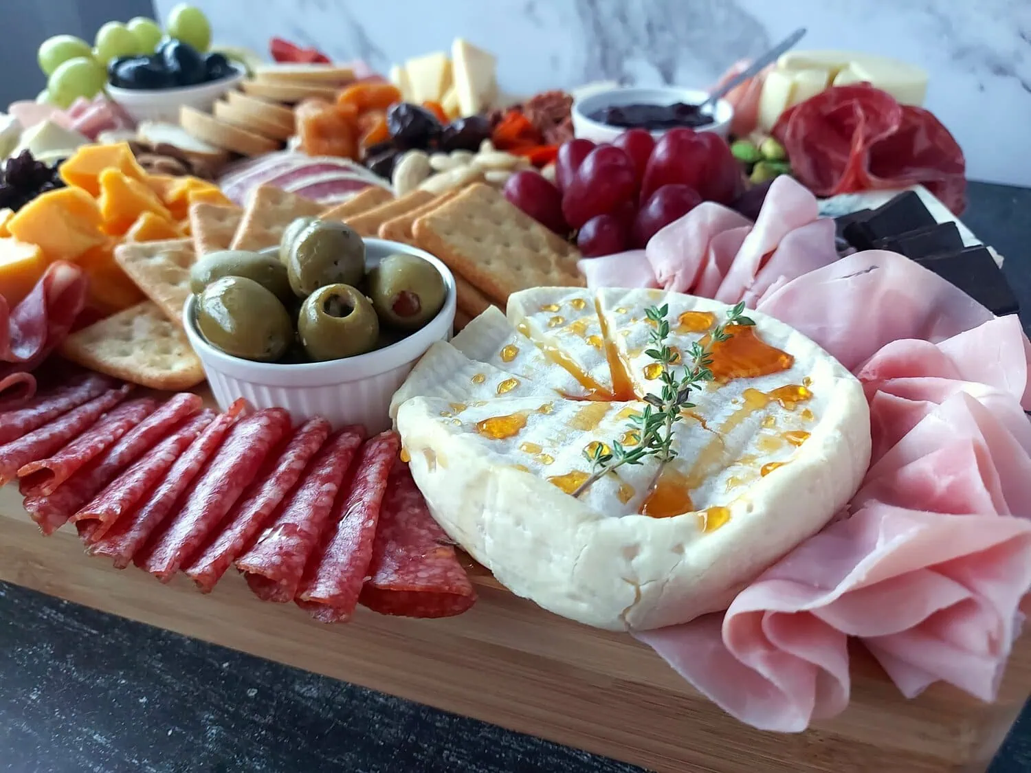 closeup of brie cheese drizzled with honey topped with a thyme sprig, next to salami, ham, olives, crackers and red grapes