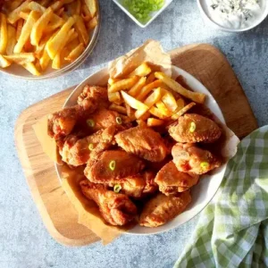 Plate full of fried chicken wings and French fries, with blue cheese dipping in the background and a plate of French fries