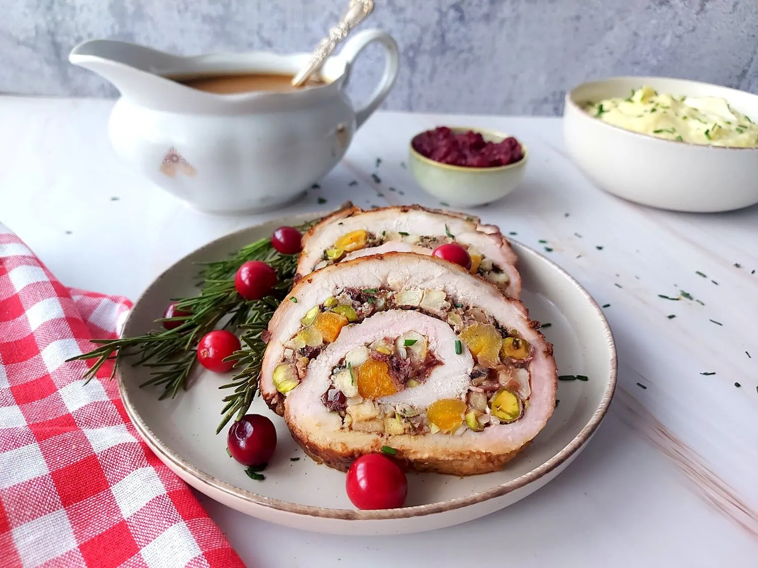 cranberry apple stuffed pork loin Three slices of stuffed pork roast on a plate with cranberries, rosemary sprig, and gravy, cranberry sauce and mashed potatoes in the background