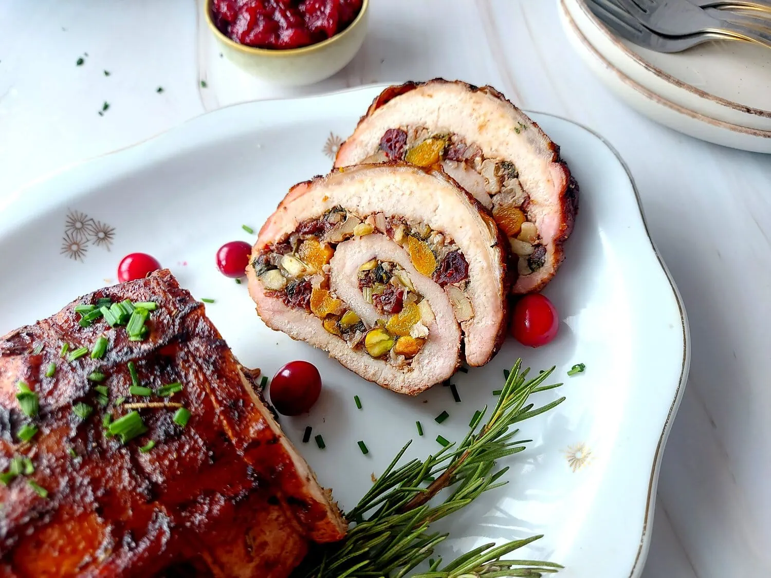 cranberry apple stuffed pork loin Overhead shot of a platter with pork roulade and two slices of stuffed pork, with some cranberries and a rosemary sprig