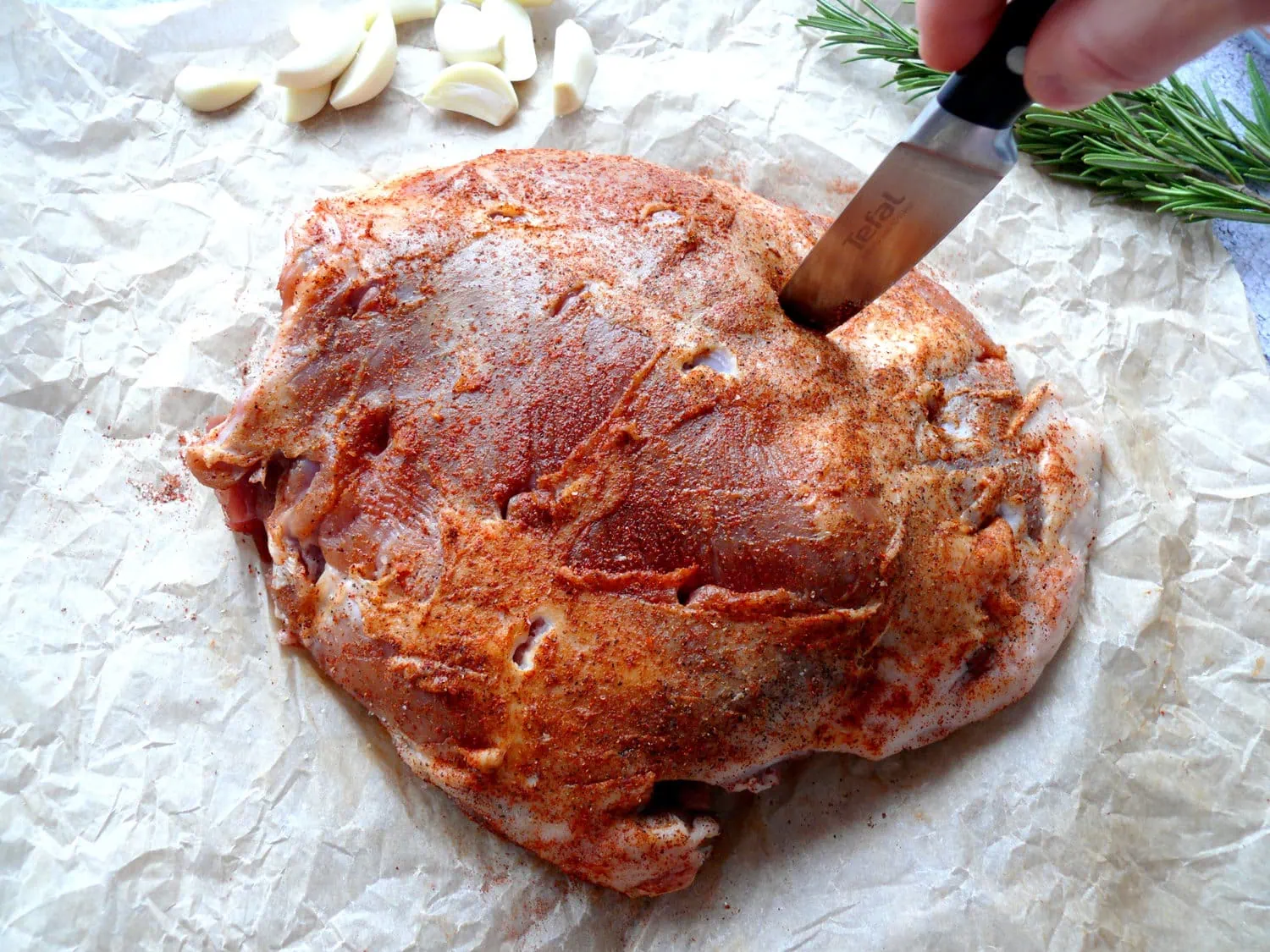 Making incisions in a raw piece of lamb, with garlic slivers and rosemary sprigs on the side