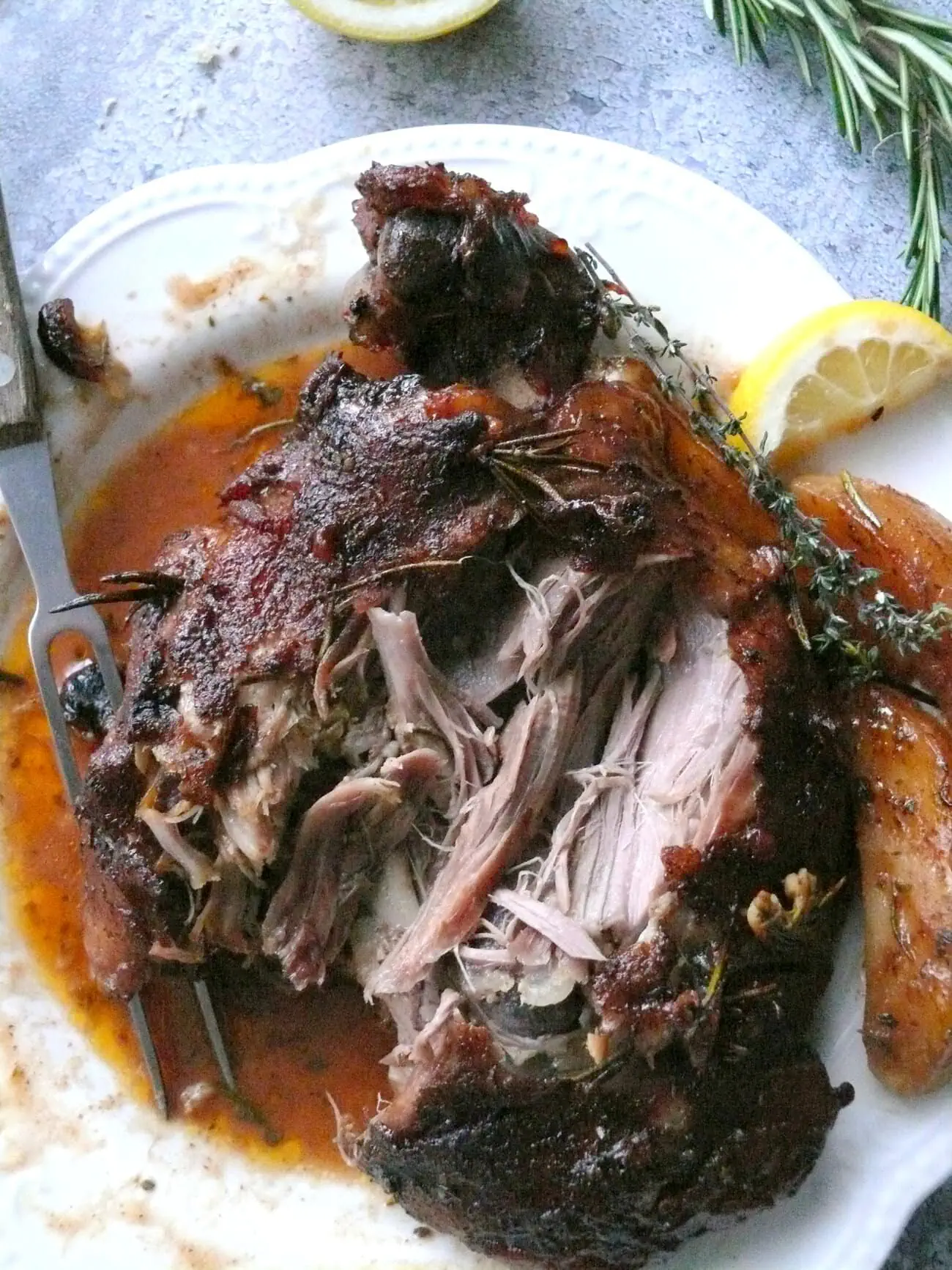 Overhead shot of a whole leg of lamb cooked in its gravy, on a white plate, shredded on the side