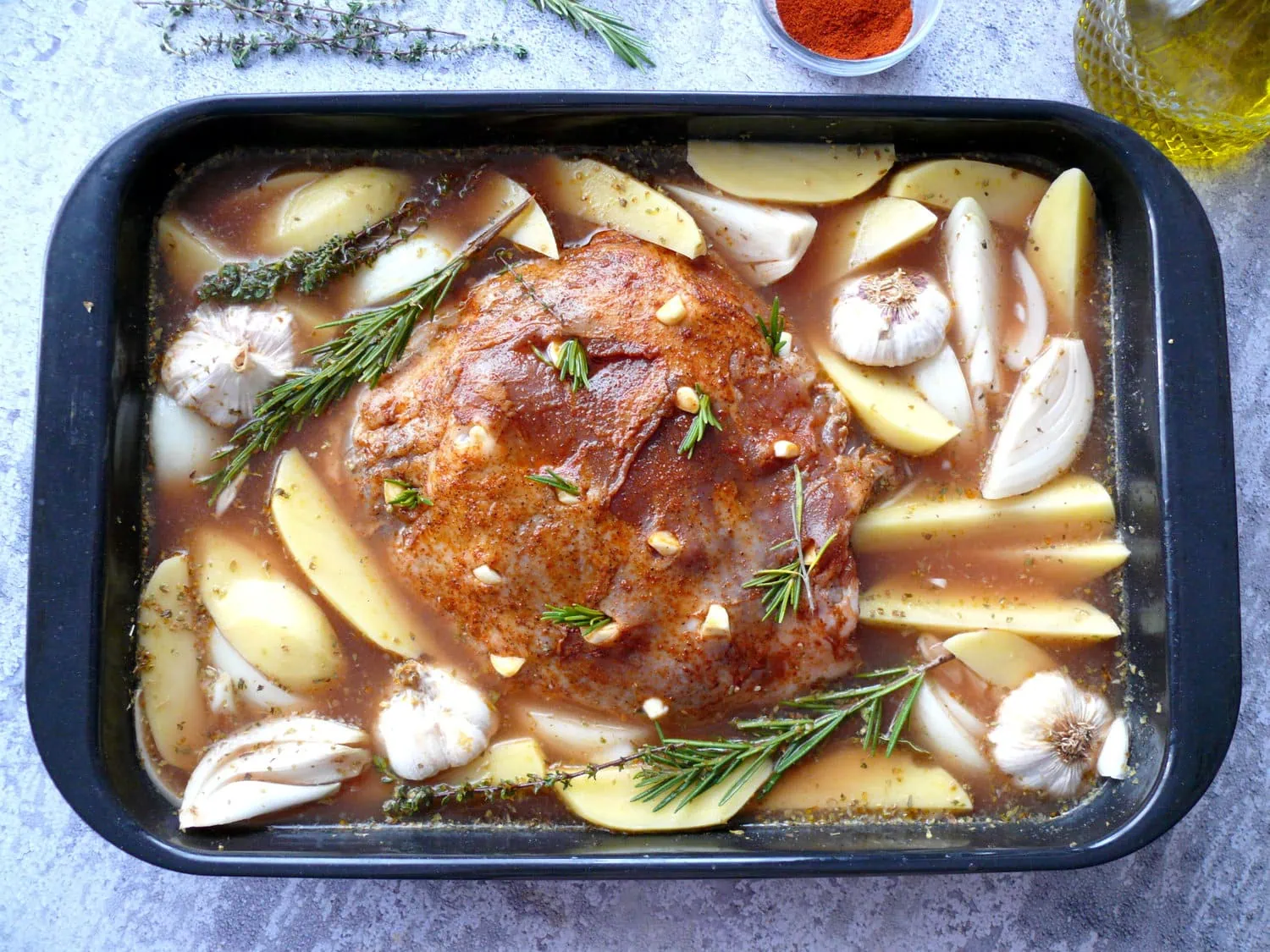 marinated lamb leg in a roasting pan over wedge potatoes, with onions, garlic, rosemary, thyme and braising liquid