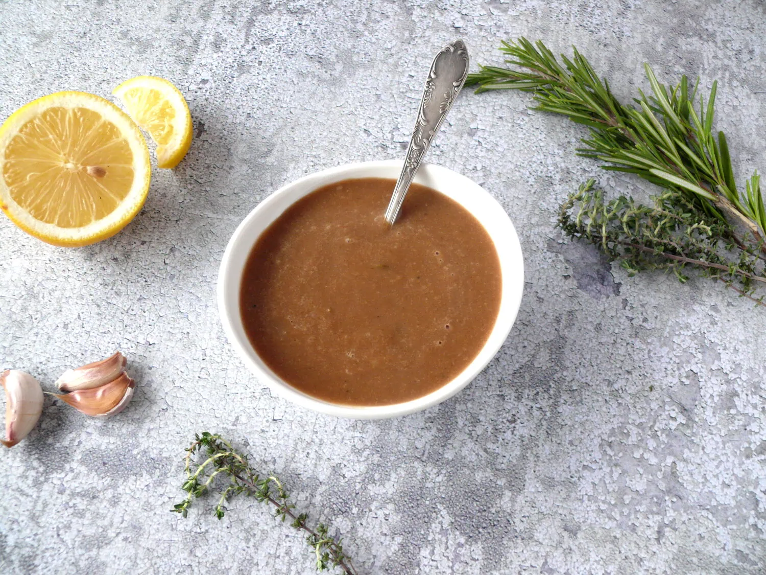 Small bowl with gravy and a spoon with lemon, garlic cloves and rosemary sprigs around