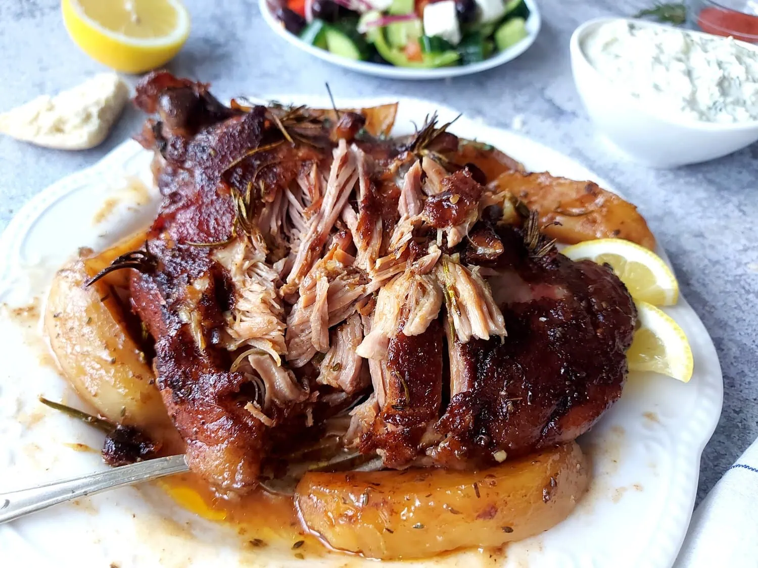Whole lamb roast partially shredded on a plate with greek lemon potatoes and gravy, with tzatziki sauce and Greek salad in the background 