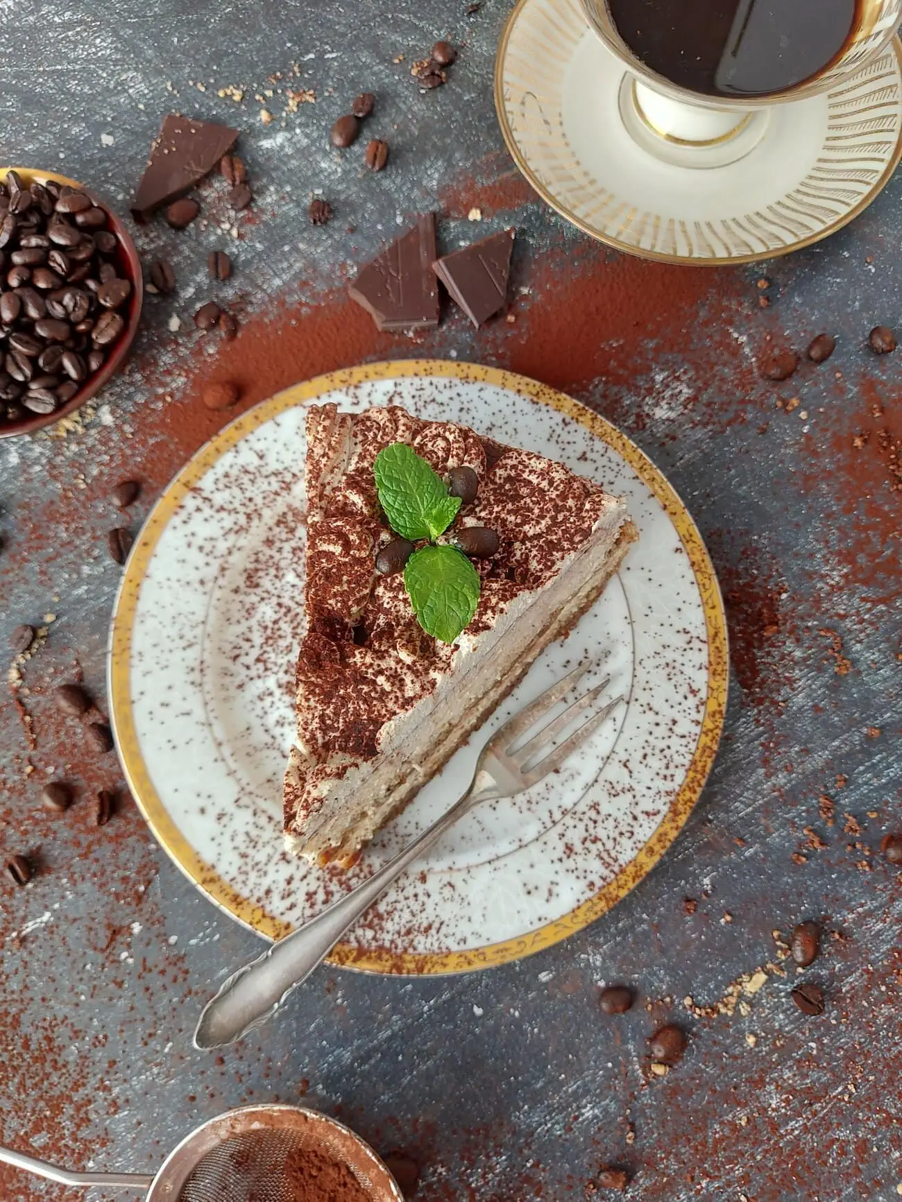 Overhead shot of a slice of cheesecake tiramisu on a white plate dusted with cocoa powder, coffee beans and dark chocolate