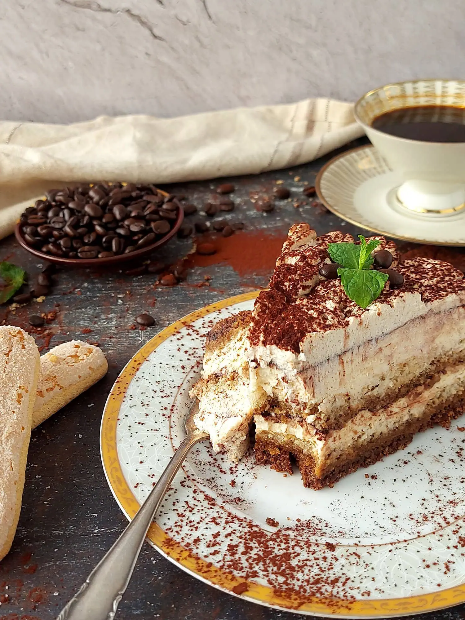 Slice of a tiramisu cheesecake on a place with a fork, and a coffee cup and lady fingers in the background