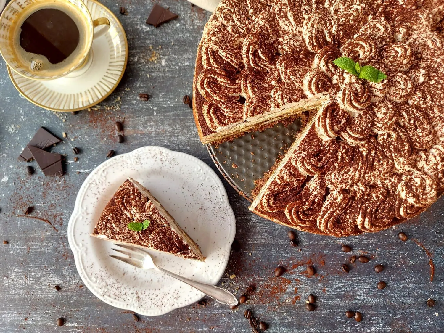 overhead shot of a slice of cheesecake tiramisu on a white plate, next to the full cake and a cup of espresso 