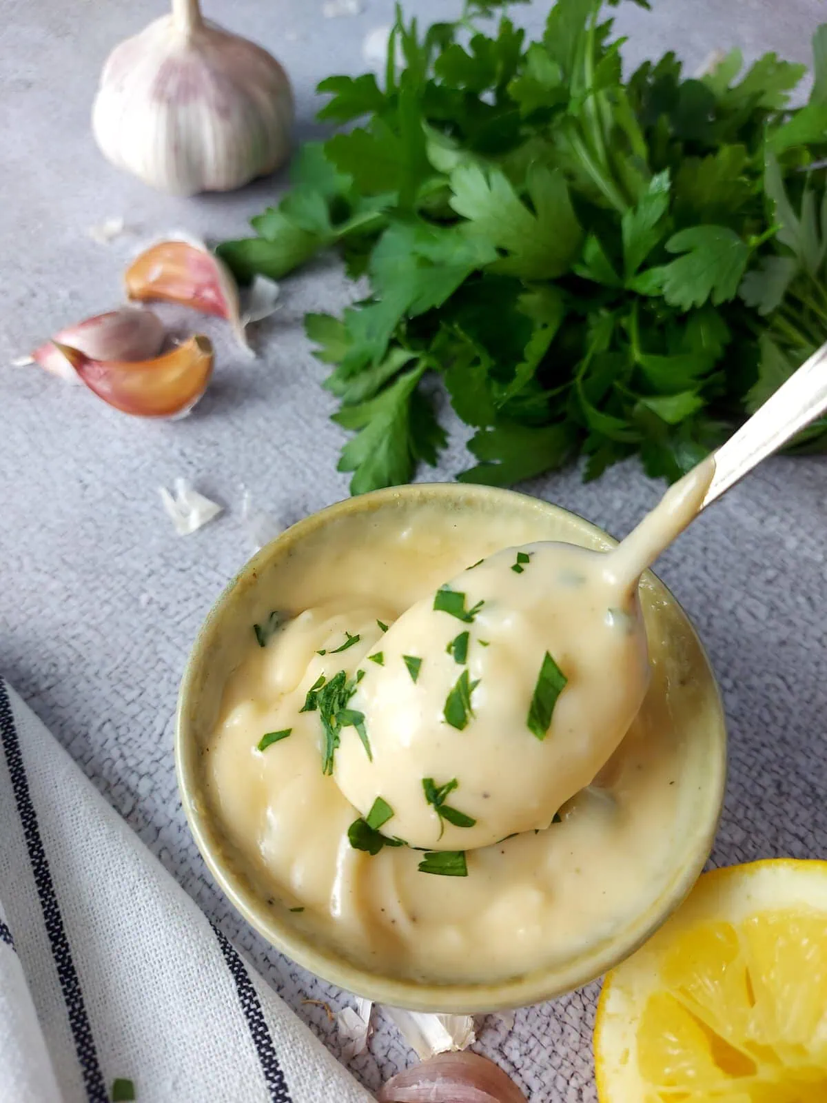 Homemade aioli with a spoon, next to a lemon, garlic cloves and a bunch of parsley